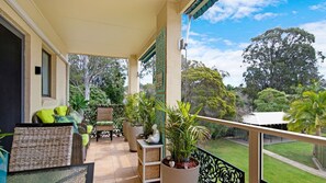 A covered balcony with wicker furniture, potted plants, and a view of a lush garden. - Rest & Play the Batehaven Way (Batehaven)