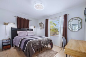A bedroom with a bed, bedside table, lamp, window with curtains, and a mirror on the wall.