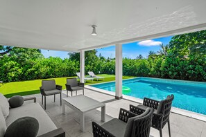 Uma área externa coberta com piscina, mobiliário e vista para um jardim bem cuidado com árvores.