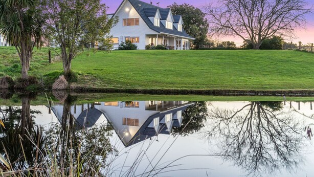 A large white house with a steep roof, surrounded by a well-maintained lawn and a body of water reflecting the house. - ⭐The Pond House by Homello⭐ | Pool • Pond • Peaceful Country Retreat (Karaka)