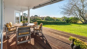A covered outdoor area with a wooden deck, a round table with chairs, and a view of a well-maintained garden and distant landscape. - ⭐The Pond House by Homello⭐ | Pool • Pond • Peaceful Country Retreat (Karaka)