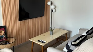 A modern living room with a flat-screen TV mounted on a wooden panel wall, a small table with a plant, a remote control, and a sofa with a throw blanket. - URBAN OASIS II (Godoy Cruz)