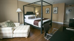 A four-poster bed with a headboard, a bedside table with a lamp, a chair, and a couch in a hotel room.
