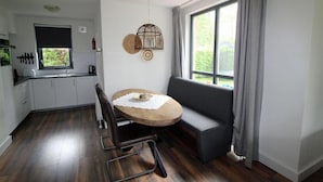 A modern kitchen with white cabinets, a dining table with a wooden top, a bench, and a large window with curtains. - Bright Bungalow in Peaceful Park Setting (Ewijk)