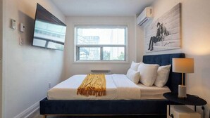 A modern bedroom with a large bed, a television mounted on the wall, a window with a view of buildings, and a painting on the wall. - Hideaway Studio heart DT MTL (Montréal)
