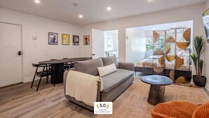 Un salon moderne avec un canapé, une table basse et un coin repas avec des chaises. Une grande fenêtre offre une vue sur l’extérieur.