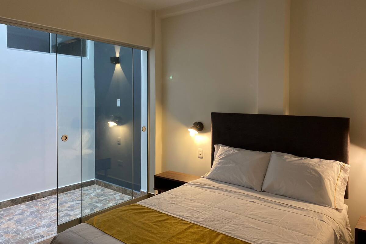 Un dormitorio con cama, mesita de noche, lámpara fijada a la pared y puerta corrediza de vidrio que da a un balcón.