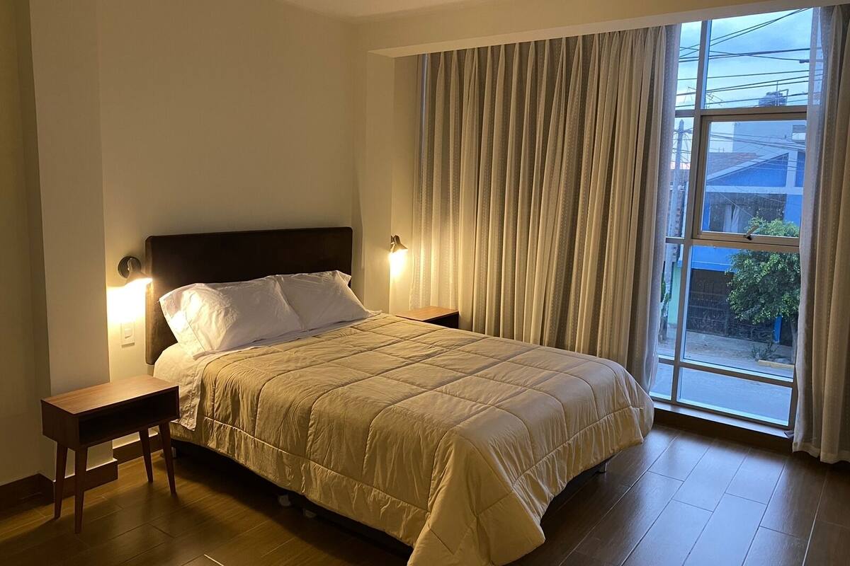 Un dormitorio con cama, mesitas de noche, ventana con cortinas y vistas al exterior.