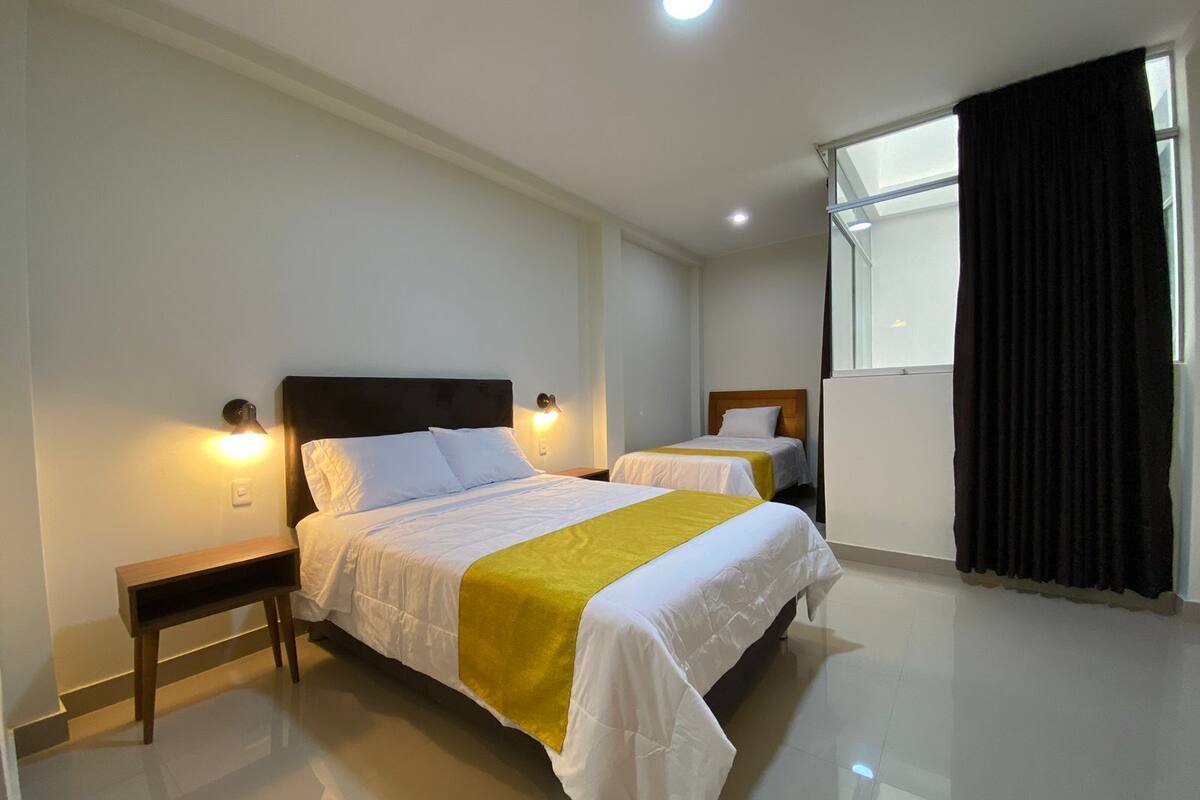 Habitación de hotel con una cama grande, dos mesitas de noche, una ventana con cortinas y una lámpara en el techo.