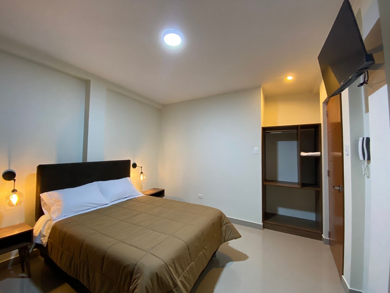 Una habitación de hotel con cama, lámparas de noche, un televisor montado en la pared y un armario.