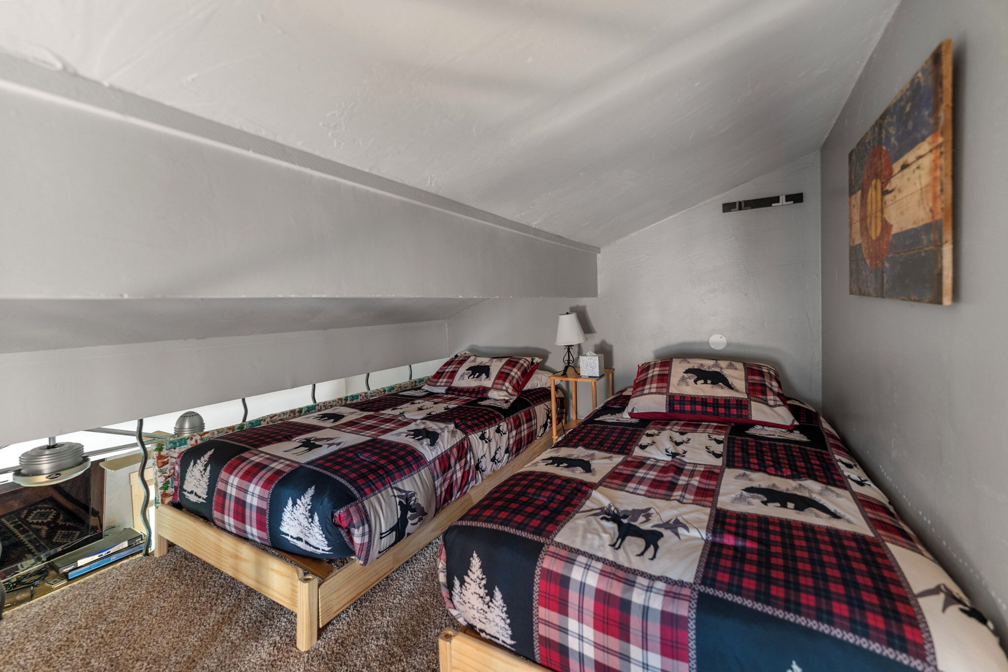 A room with two wooden beds, each with plaid bedding featuring animal prints. There is a small table with a lamp and a wall-mounted shelf.
