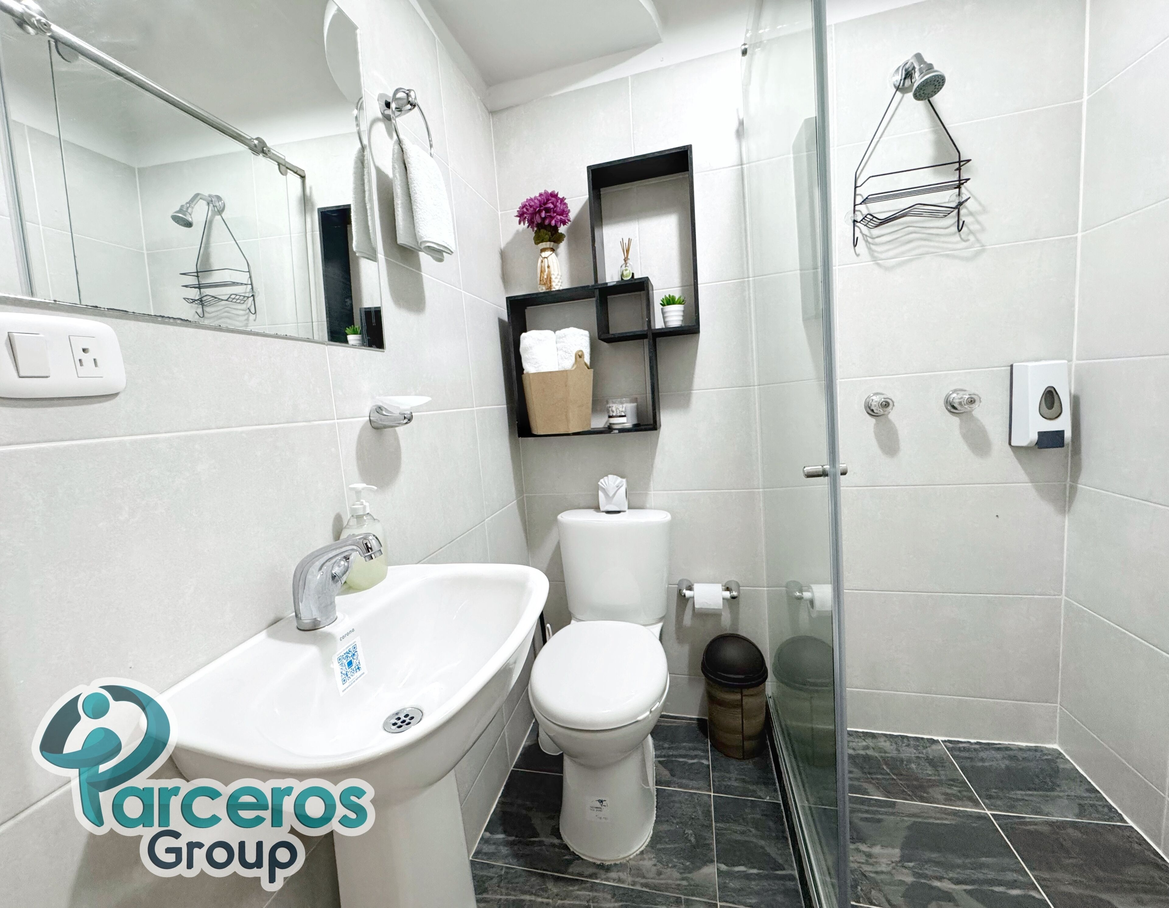 A modern bathroom with a white sink, toilet, and shower. There are shelves with toiletries and a glass shower enclosure.