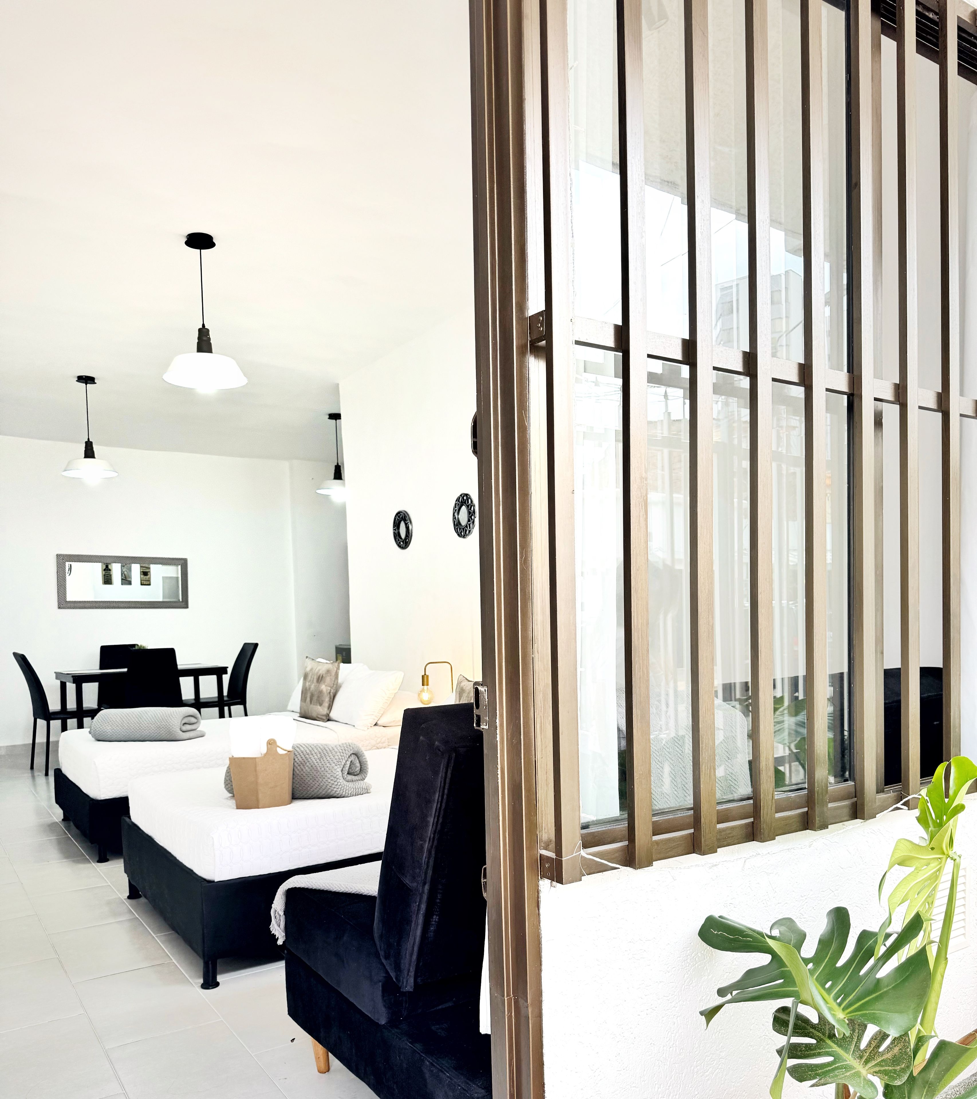 Habitación de hotel moderna con cama, escritorio y silla. Se aprecia un ventanal grande con persianas verticales y una planta en maceta sobre el alféizar.