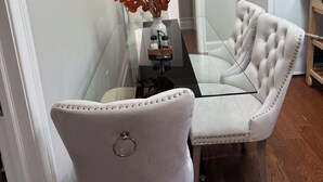 Un comedor con una mesa de vidrio, un sillón capitonado, un jarrón con flores secas, un refrigerador y un microondas.