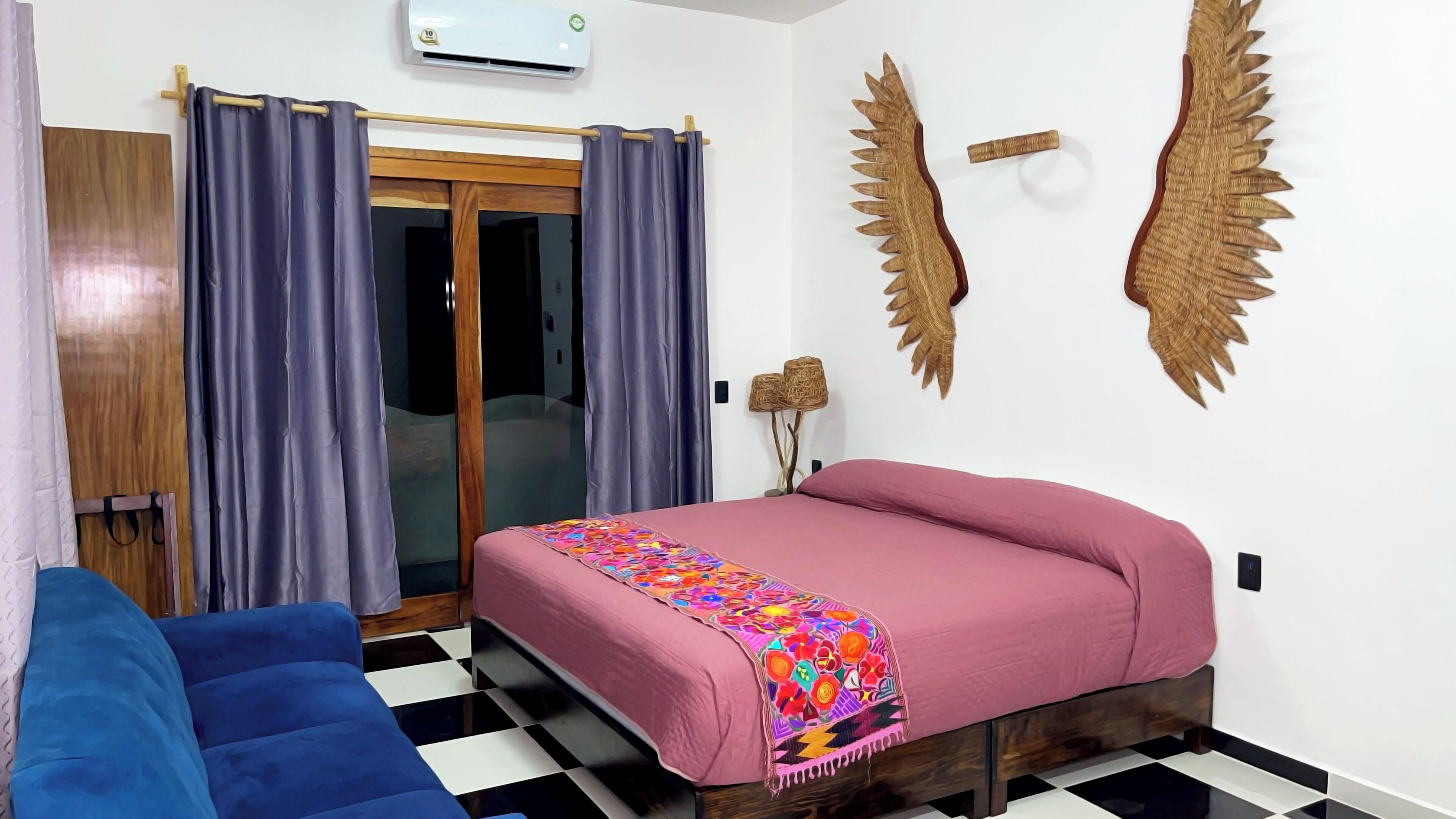 Quarto com cama, sofá azul, piso xadrez, porta de madeira e ar-condicionado fixado na parede.