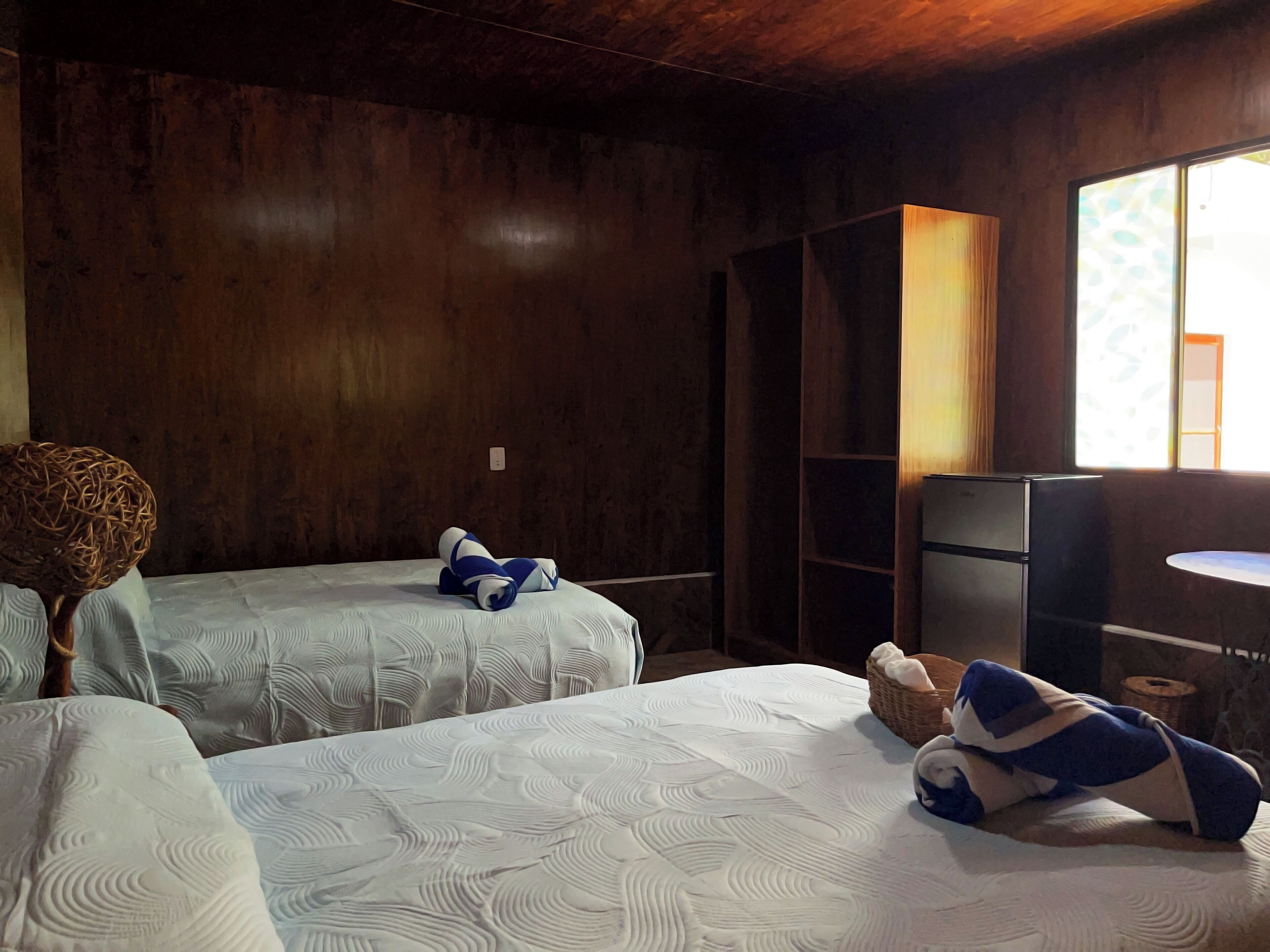 A room with two beds, wooden walls, a small refrigerator, and a window with natural light.