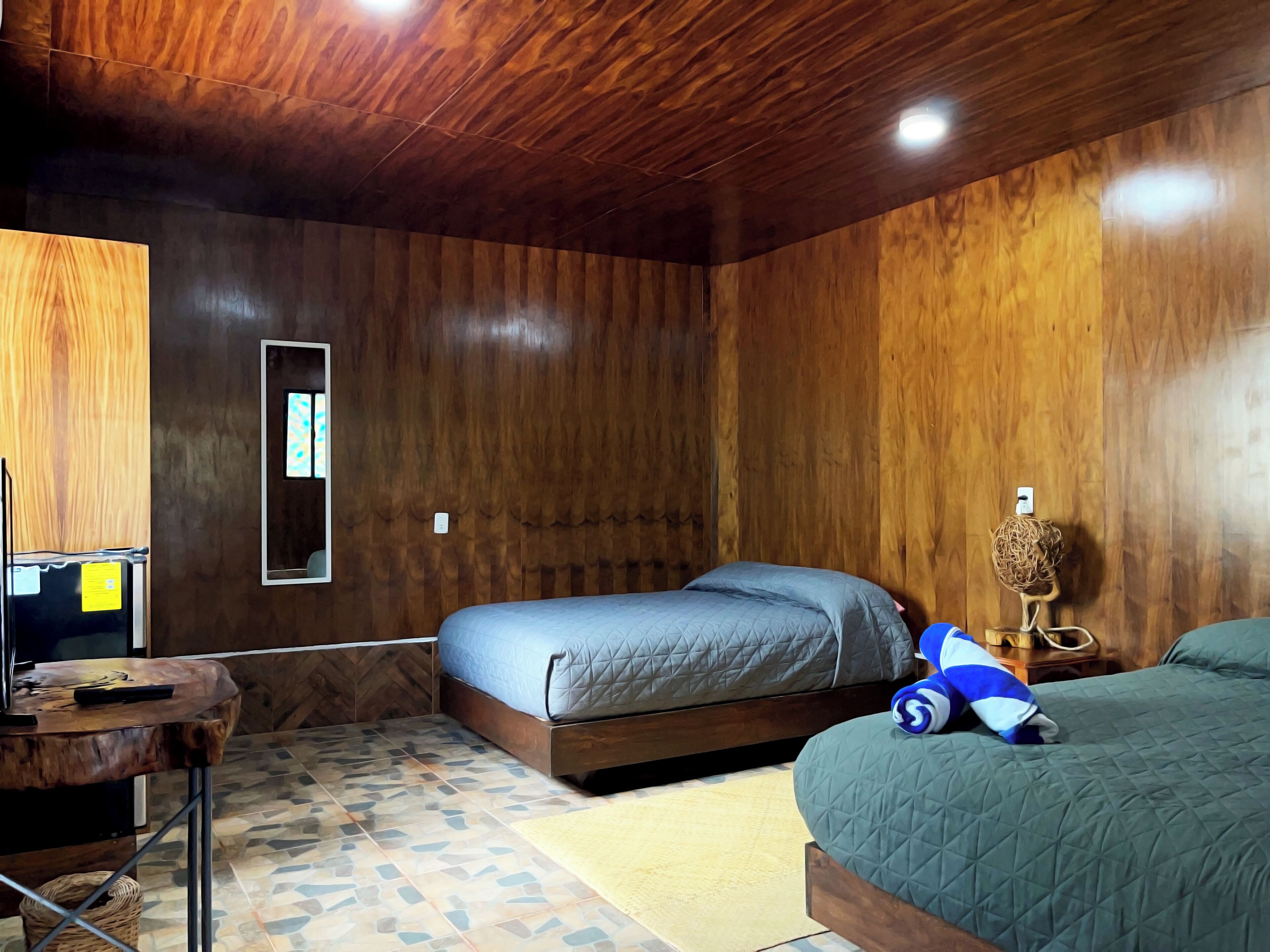 Una habitación con techo de madera, cama con colcha azul, una mesita con un control remoto y un objeto decorativo en la mesita de noche.