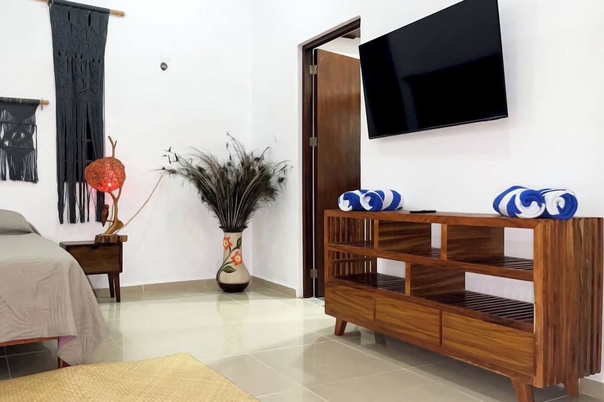 A modern bedroom with a bed, a wooden side table, a TV mounted on the wall, and a decorative vase with peacock feathers.
