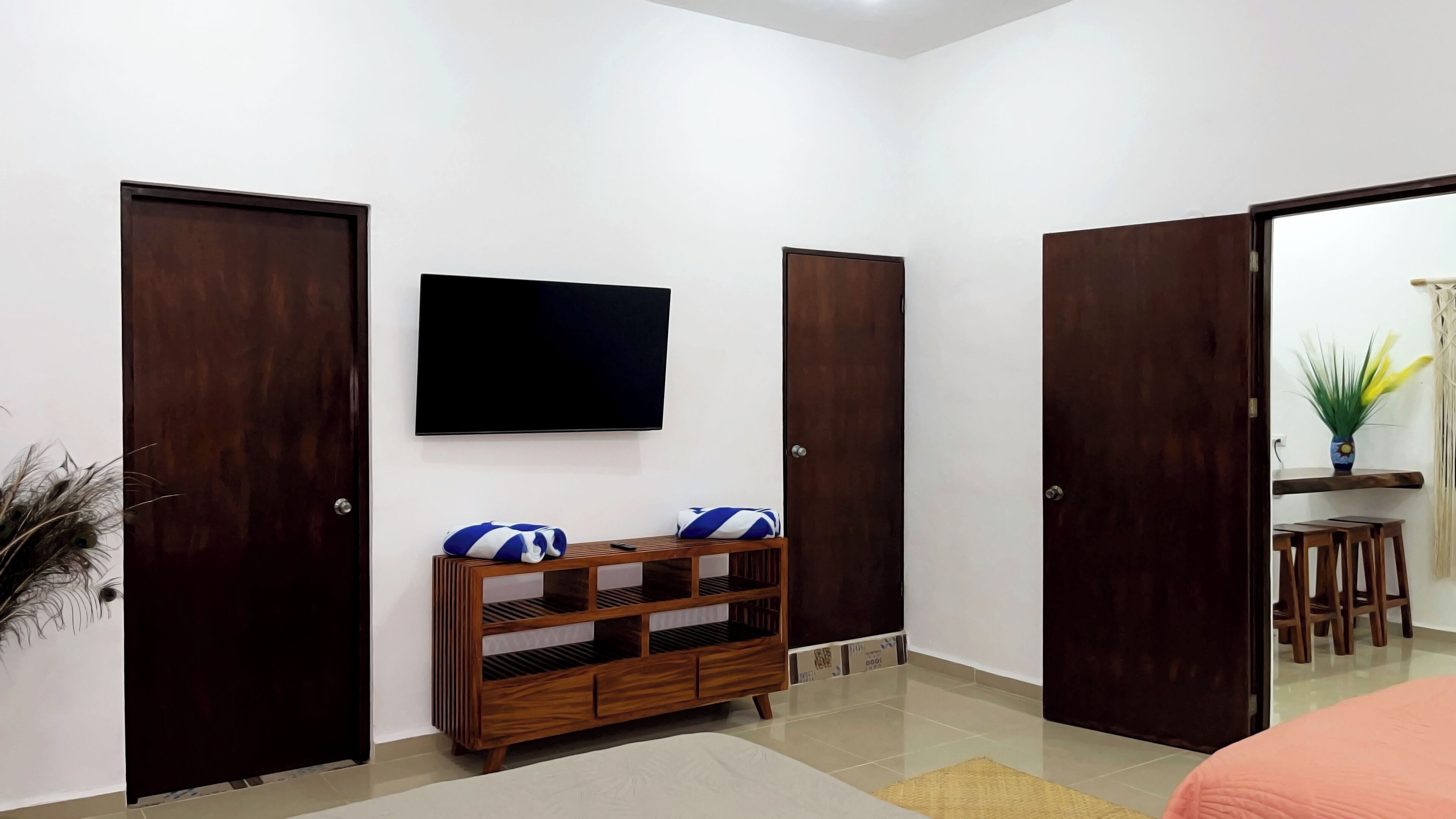 Habitación de hotel con un televisor montado en la pared, una mesita de madera con toallas, y dos puertas.
