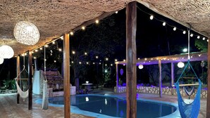 Zona de piscina al aire libre con hamacas colgantes, luces de cadena y un muro de piedra de fondo.