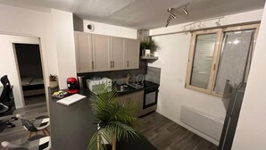 A compact kitchen with a black countertop, white cabinets, and a built-in oven. There is a small island with a plant, and a window with blinds. - Apartment le Bugatti 4 persons wifi (Yvré-l'Évêque)