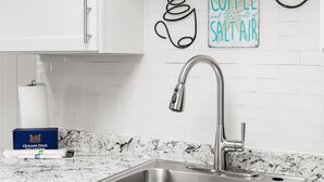 Eine Küche mit weißen Schränken, einer Arbeitsplatte aus Granit, einer Spüle aus Edelstahl und einem Schild an der Wand, auf dem steht: 'all good is coffee and the smell of salt air.'