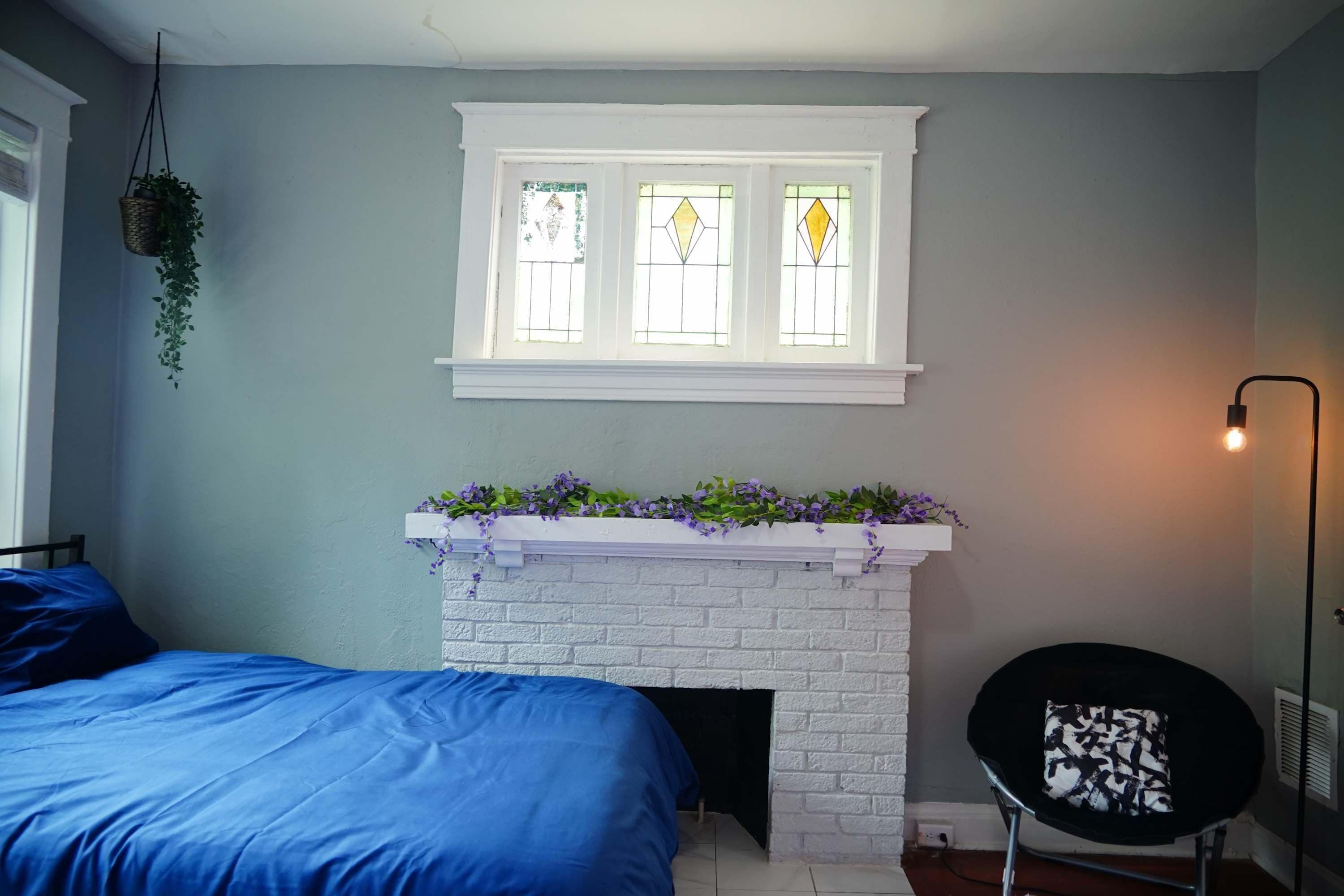 Quarto com cama azul, lareira de tijolos brancos e planta pendente.
