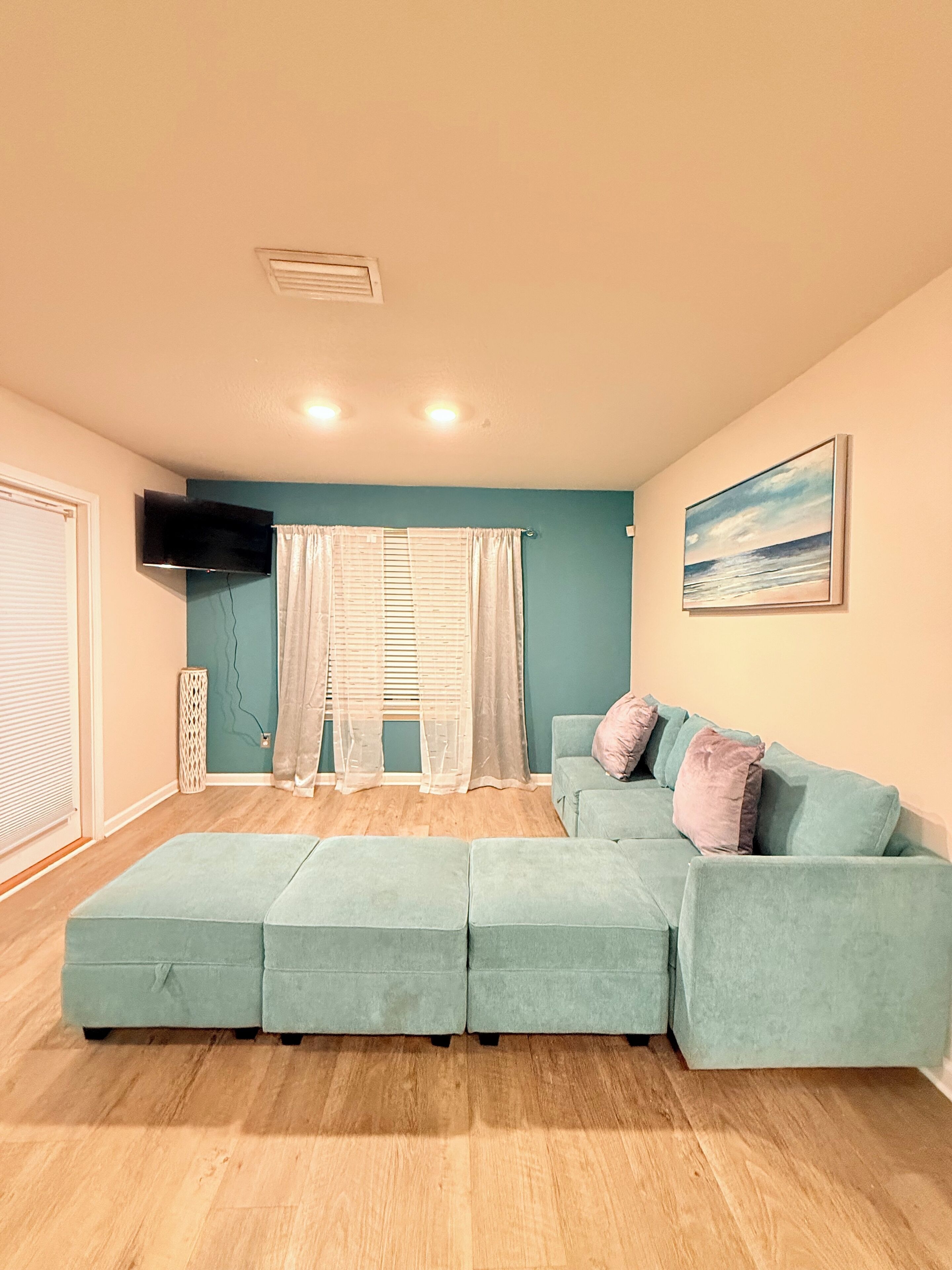 A living room with a sectional sofa, a flat-screen TV mounted on the wall, and a painting of a beach scene.