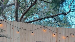 Un jardin avec une clôture en bois, un barbecue, des guirlandes lumineuses et des plantes en pot.
