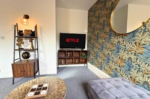 A living room with a television displaying 'NETFLIX', a wooden cabinet, a globe, a shelf with decorative items, a round coffee table, and a patterned wallpaper. - Smart 3-Bed in Hexham-Ideal for Workers & Families (Hexham)
