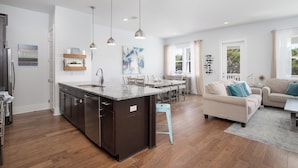 Een moderne keuken met donkere kasten, een granieten aanrecht en hanglampen. Er is een eethoek met een witte tafel en stoelen, en een beige bank in de woonkamer.