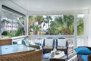 A covered outdoor seating area with wicker furniture, a glass table, and a view of palm trees and a building in the background.
