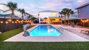 Un patio trasero con piscina, mobiliario de exterior e iluminación colgante.