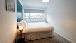 Une chambre à coucher avec un lit, une table de chevet, une lampe et une fenêtre donnant sur les bâtiments environnants.