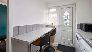 Une cuisine compacte avec un plan de travail blanc, deux tabourets de bar noirs, un four à micro-ondes et une porte ornée d’un vitrail.