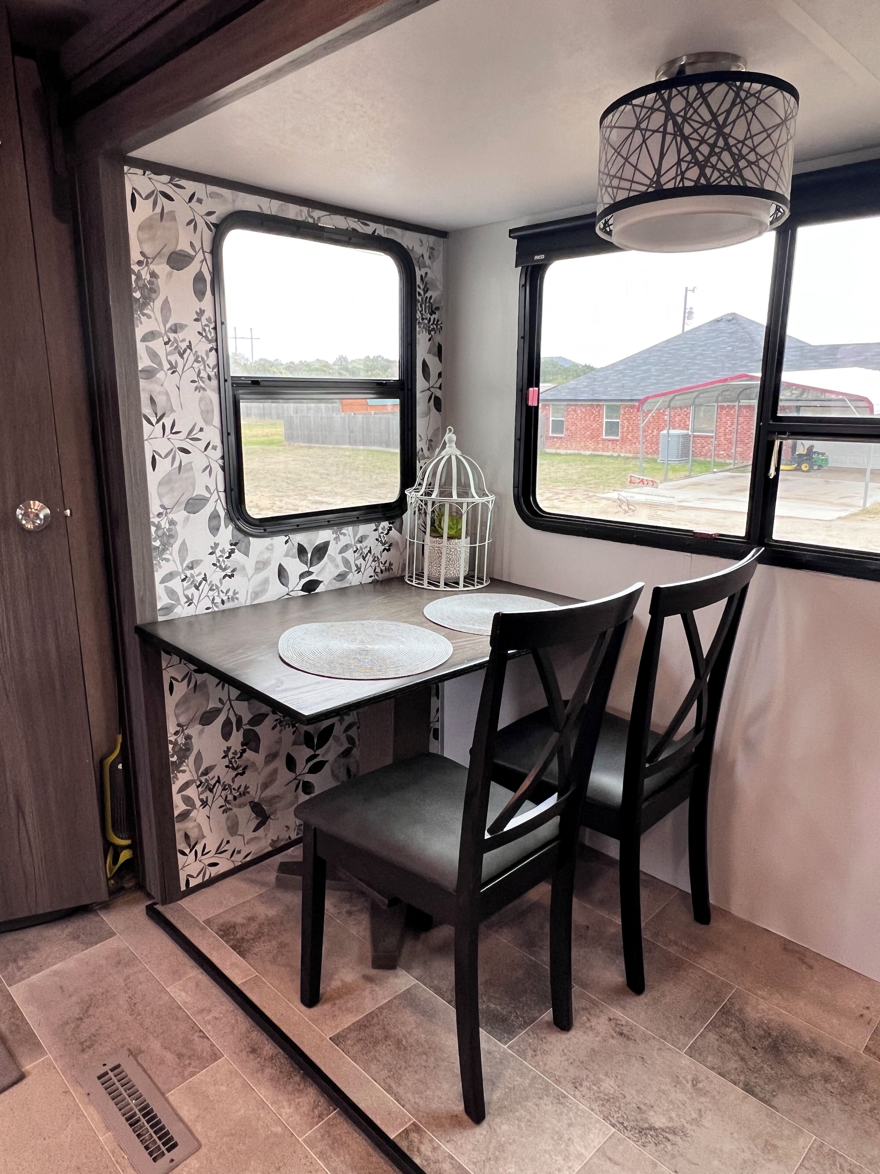 Una cocina compacta con una mesa de comedor, dos sillas, un ventanal con vistas a un edificio de ladrillo y una jaula decorativa en el alféizar.
