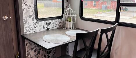 Una cocina compacta con una mesa de comedor, dos sillas, un ventanal con vistas a un edificio de ladrillo y una jaula decorativa en el alféizar.