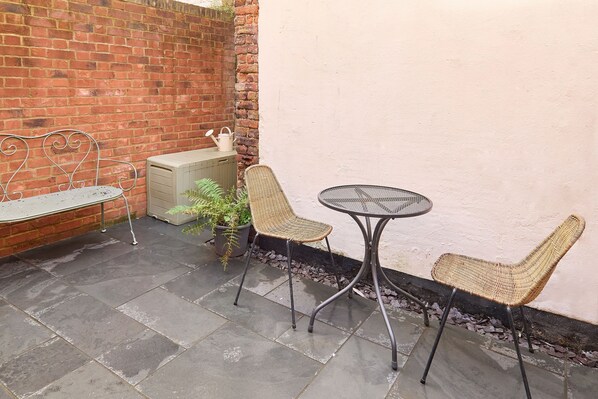 A patio area with a brick wall, a metal bench, a wicker chair, a round table, and a potted plant. - Host Stay Aurora (Deal)