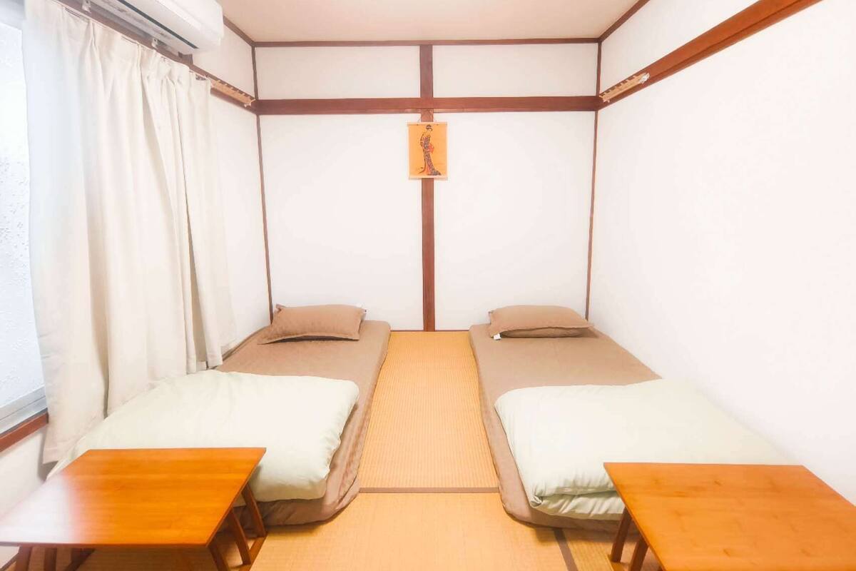 A small, single-room hotel room with two single beds, a wooden table, and a window with curtains.