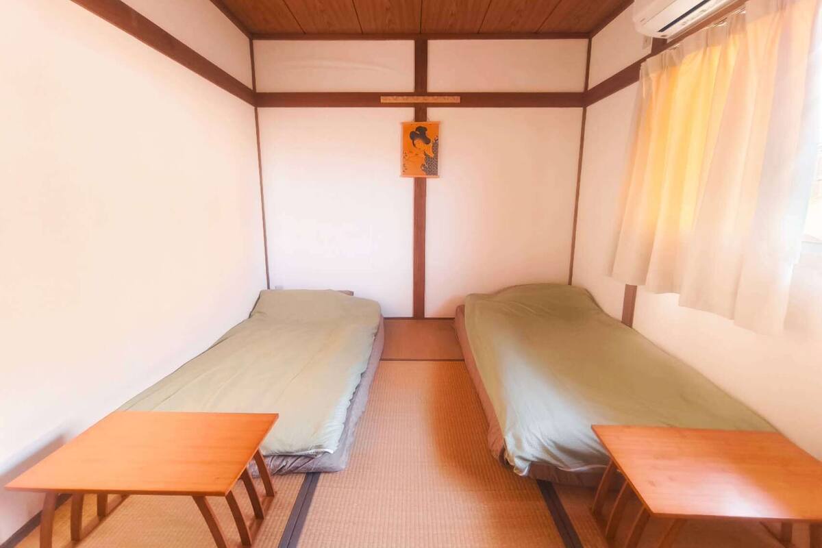 A small, simple room with two single beds, a wooden table, and a futon.