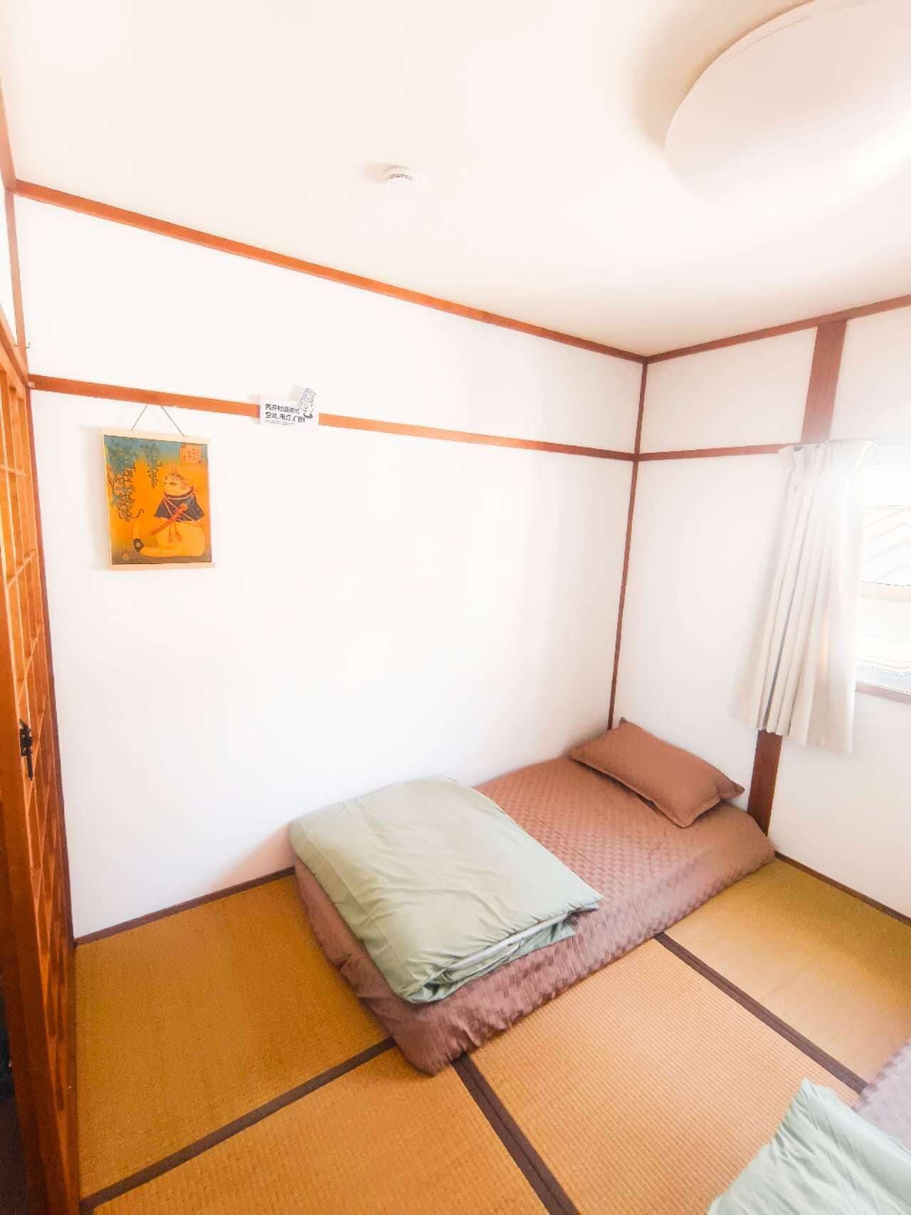 A small, single-bed room with tatami flooring, a simple bed with a green and brown blanket, a small painting on the wall, and a window with curtains.