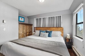 Ein Schlafzimmer mit einem großen Bett, einem Holz-Kopfteil, einem Fenster mit Vorhängen und einem kleinen Nachttisch mit Taschentuchbox.