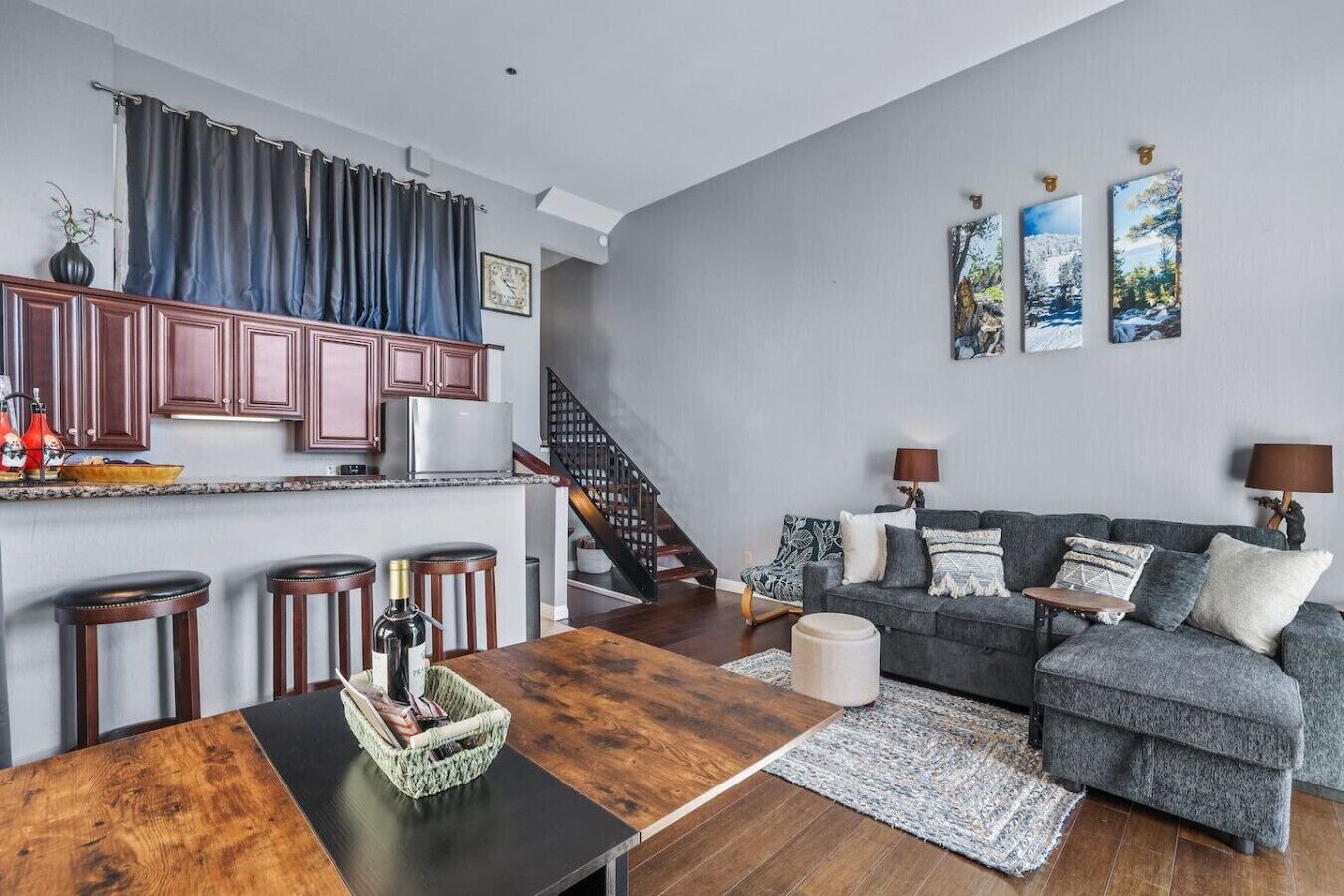 A modern living room with a grey sofa, a wooden coffee table, and a kitchen area with dark wooden cabinets and a grey countertop.