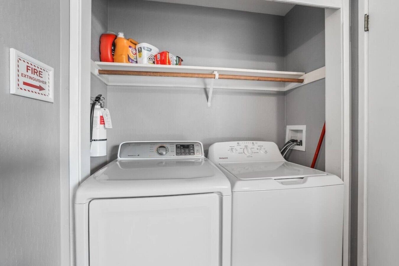 A laundry room with two washing machines, a fire extinguisher, and a shelf with cleaning supplies.