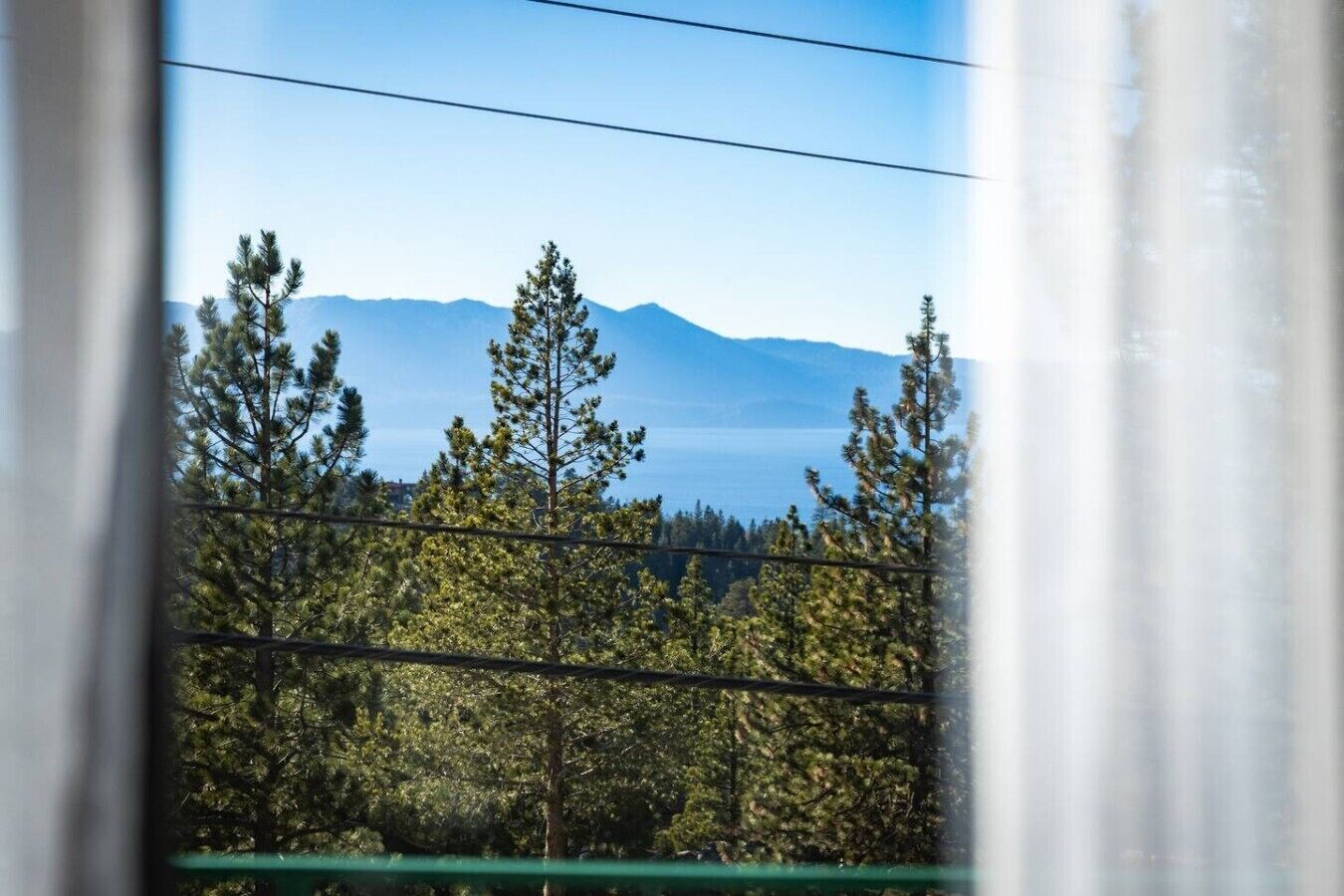Une vue à travers une fenêtre, avec une chaîne de montagnes en arrière-plan, des arbres au premier plan et des lignes électriques au-dessus.