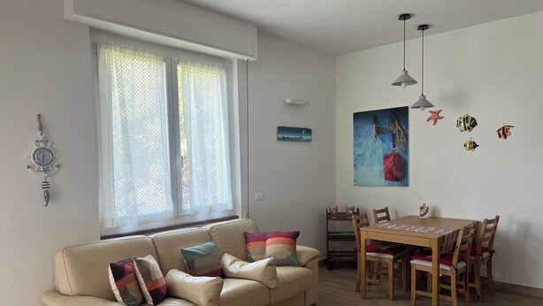 A living room with a beige sofa, a dining table with chairs, a window with curtains, a wall clock, and a painting. - Seaside Escape With Balcony Views (Portovenere)