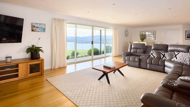 A spacious living room with a large window offering a scenic view, a leather sofa, a wooden coffee table, and a TV mounted on the wall. - Bruny Horizon - Where the Country meets the Sea (North Bruny)