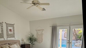 A bedroom with a bed, a ceiling fan, a nightstand, a macrame wall hanging, and a view of an outdoor pool through a glass door. - Luxury Desert 3BR house with Private Pool & Spa (La Quinta)