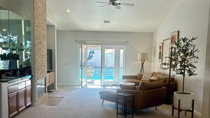 A spacious living room with a sofa, coffee table, and a potted plant. There is a kitchen area with cabinets and a countertop visible. - Luxury Desert 3BR house with Private Pool & Spa (La Quinta)