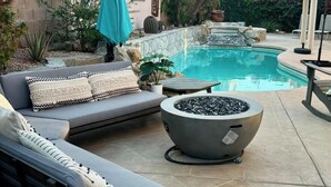A poolside area with a grey sofa, a fire pit, and a red umbrella. - Luxury Desert 3BR house with Private Pool & Spa (La Quinta)